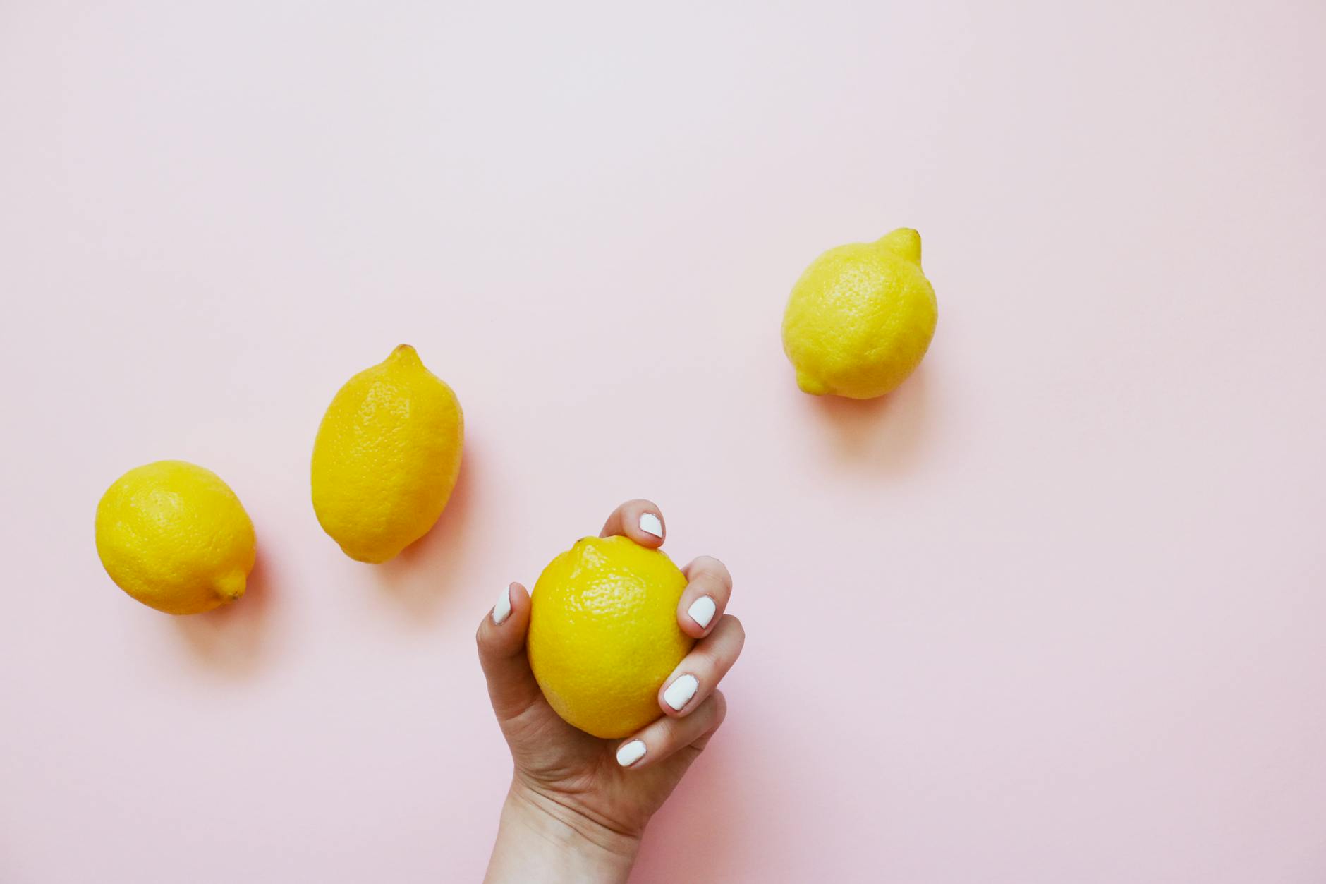 Uma mão segurando um limão amarelo em fundo rosa suave, cercada por três limões adicionais