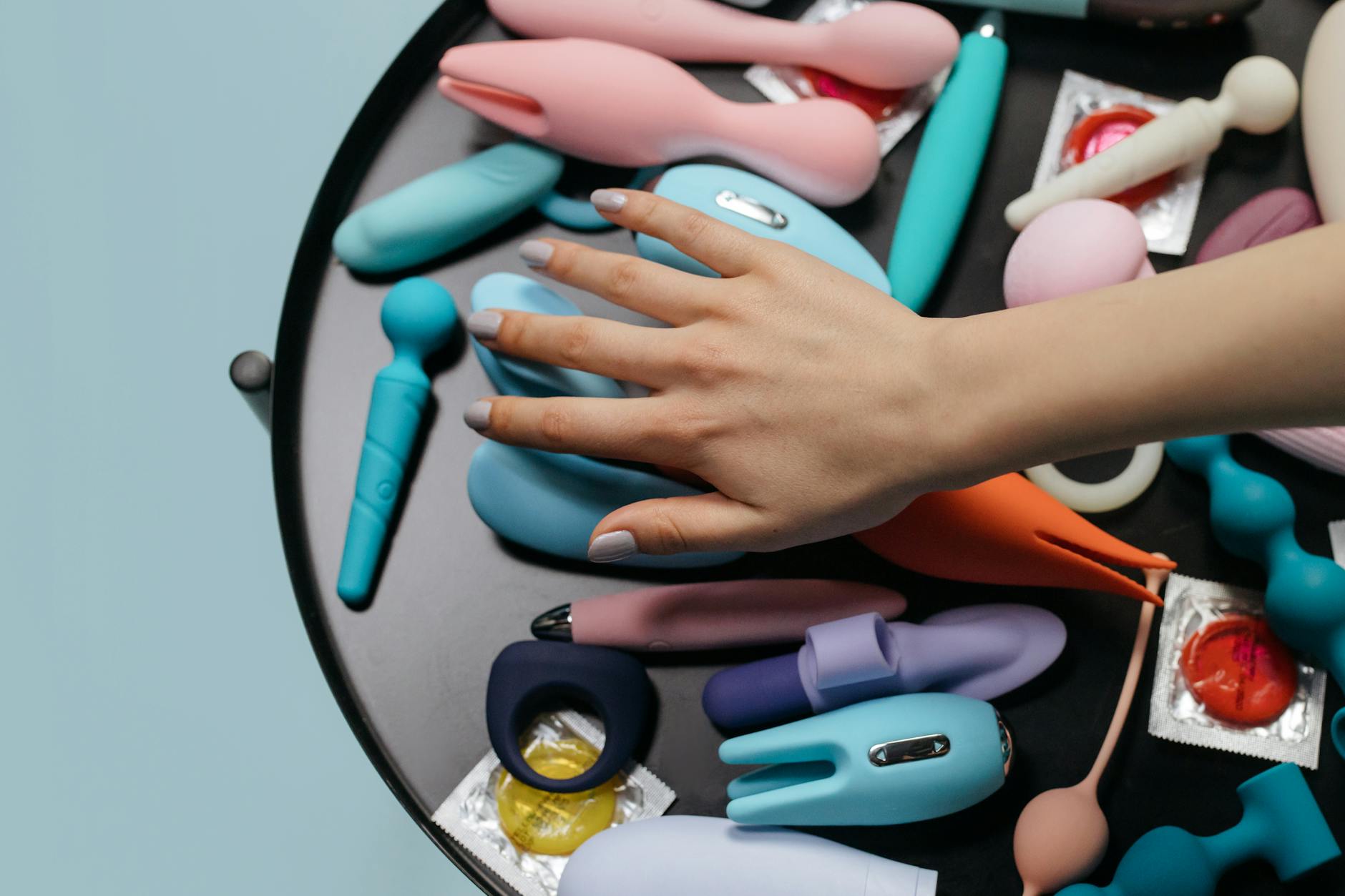 Mão estendida sobre uma variedade de brinquedos sexuais coloridos arranjados numa mesa