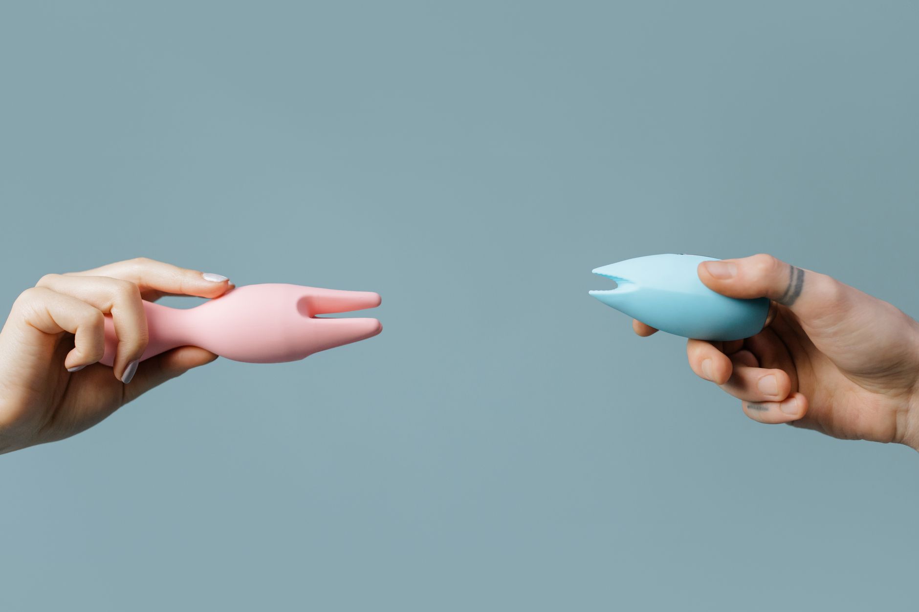 Duas mãos segurando vibradores de silicone rosa e azul contra um fundo pastel
