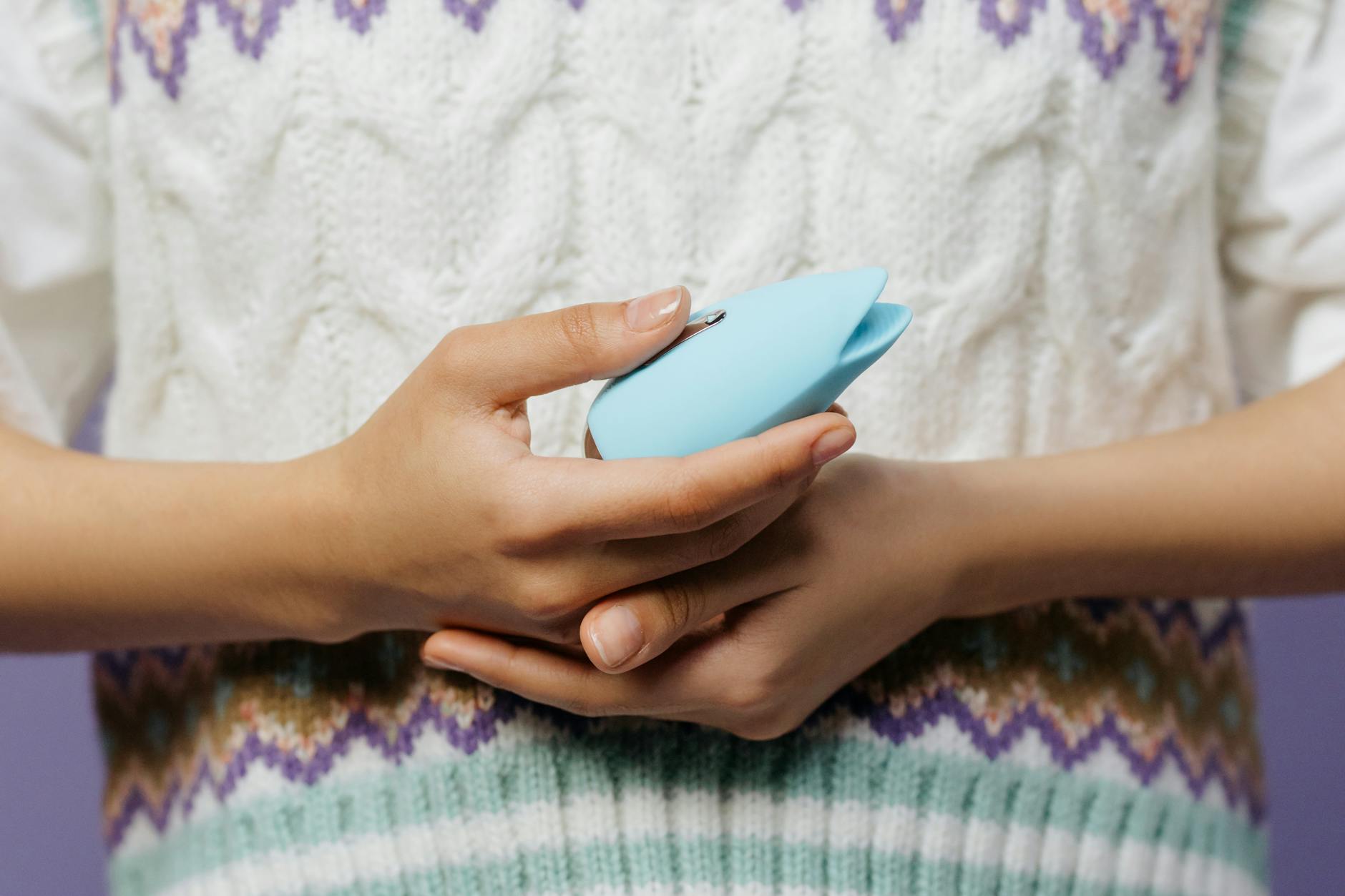 Mãos segurando um vibrador de sucção azul contra um suéter tricotado, representando conforto e prazer acessível