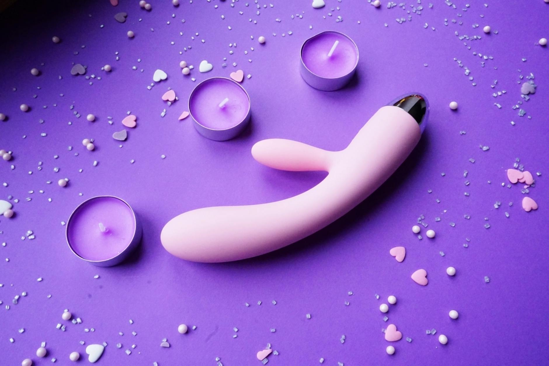 Vibrador rosa em fundo roxo com confete de corações e velas para criar clima romântico
